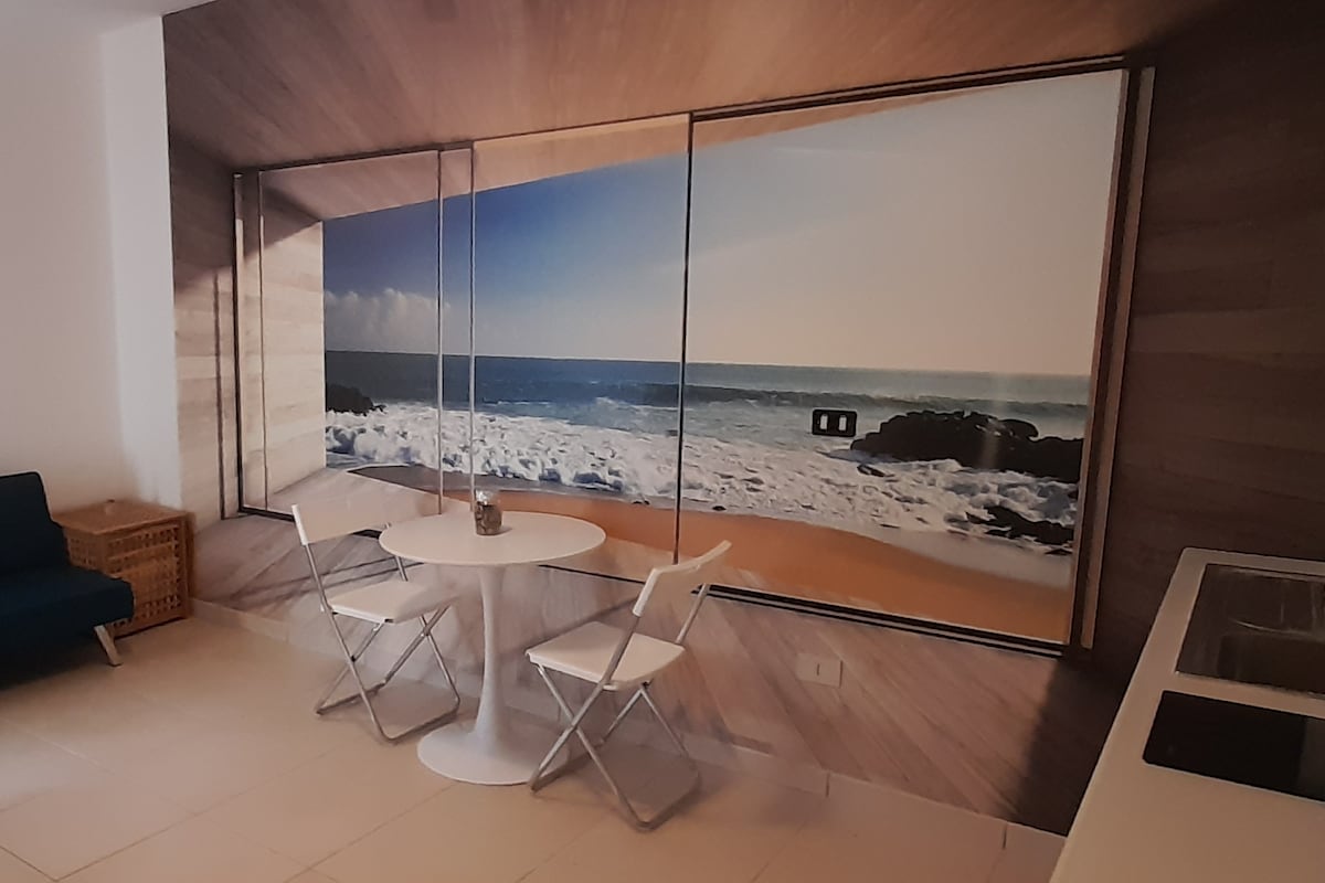 A light and airy dining area features a round table surrounded by two white chairs. A large mural of a beach scene with waves rolling onto the shore creates a serene ambiance, while a compact kitchen area is visible in the background.