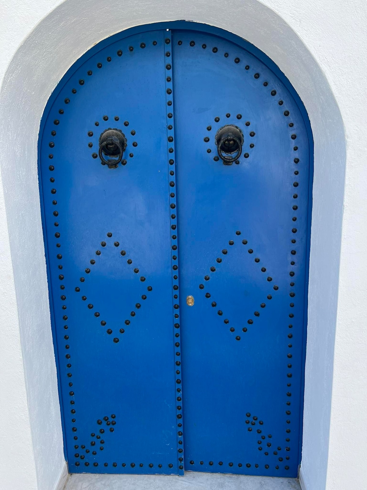 A pair of arched doors painted in a vibrant blue features decorative studs arranged in diamond patterns. Two ornate door knockers are positioned at the center of each door, set against a pristine white wall.