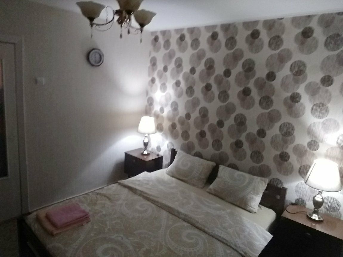 A sleeping area features a spacious bed with a patterned duvet and two pillows. Nightstands with lamps flank either side of the bed, providing soft lighting. A clock is mounted on the wall, and a decorative wallpaper adds visual interest.