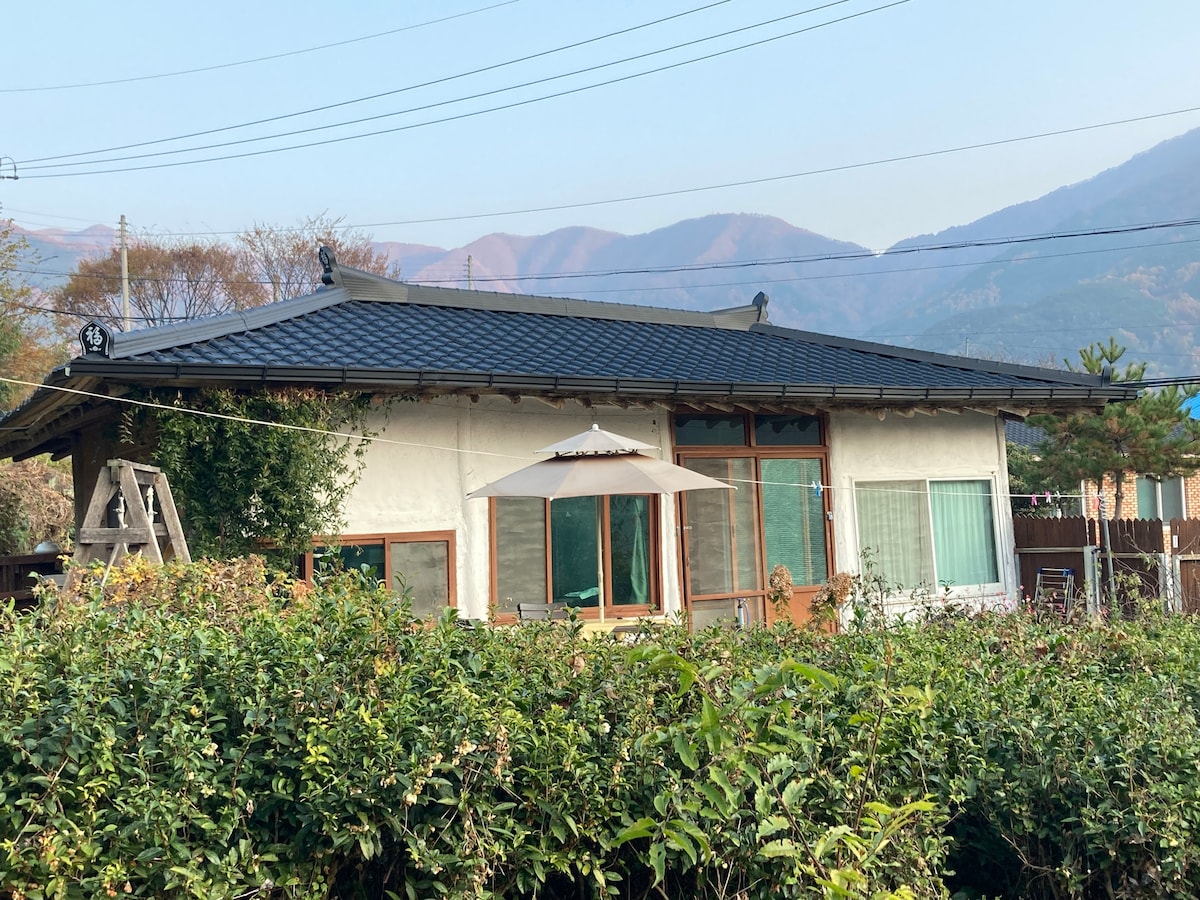A traditional Korean house, constructed from earthen materials, features a sloping roof and large windows. The exterior is surrounded by lush greenery, with mountains visible in the background, creating a serene countryside setting.