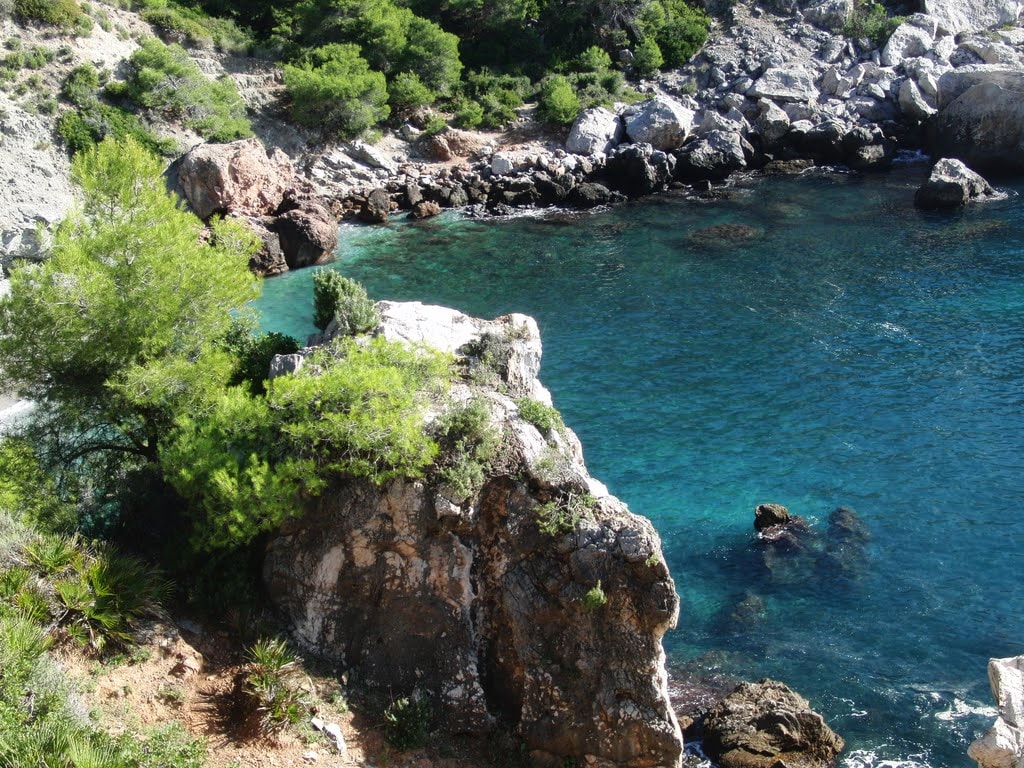 A serene coastal scene presents clear turquoise waters gently lapping against rocky shores. Lush greenery is visible among the rocks, enhancing the natural beauty of the landscape. The interplay of light creates a shimmering effect on the water's surface.