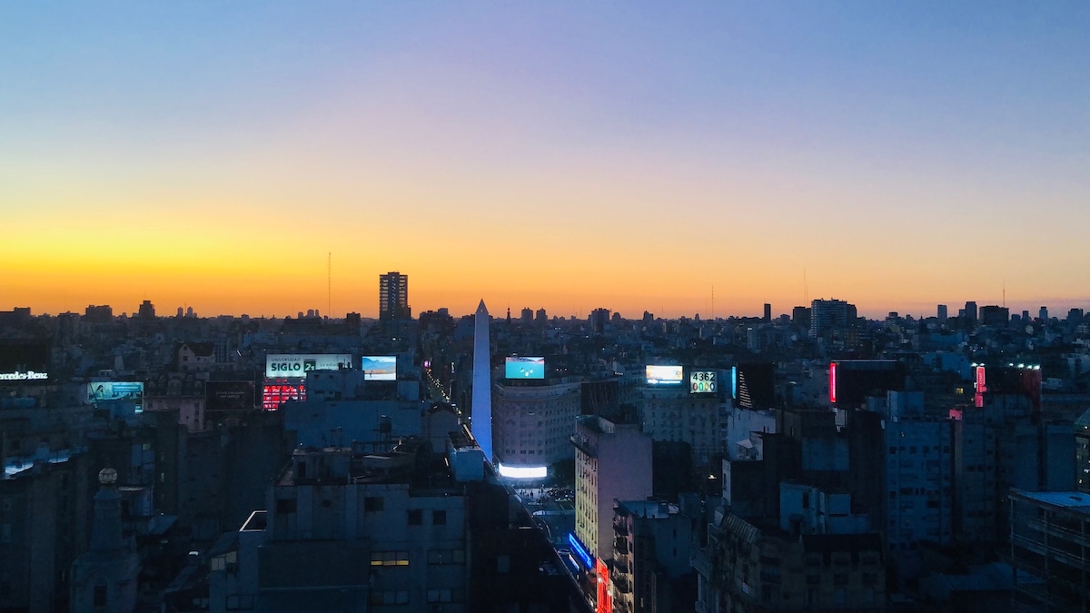The cityscape features a skyline at sunset, with a gradient of warm colors in the sky. Buildings are illuminated, showcasing various advertisements and signs. The view encompasses a mix of architectural styles, contributing to the vibrant urban atmosphere visible from the 20th floor.