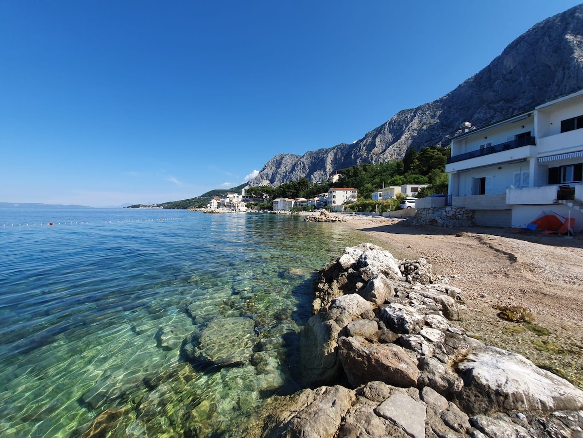 A clear view of the calm, shallow waters along a pebble beach is provided, with gently lapping waves against a rocky shore. In the background, a quaint village is nestled among green hills, and the dramatic mountains rise sharply against the clear blue sky.