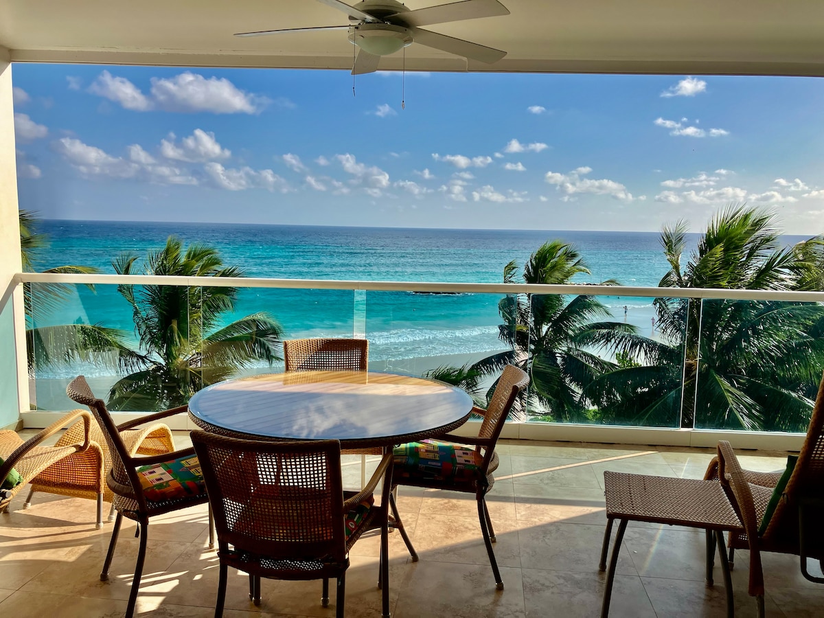 A spacious balcony with a circular table and several woven chairs offers a view of the turquoise sea and beach below. Lush palm trees sway gently in the breeze. The open space is well-lit by natural sunlight filtering through the glass railing.