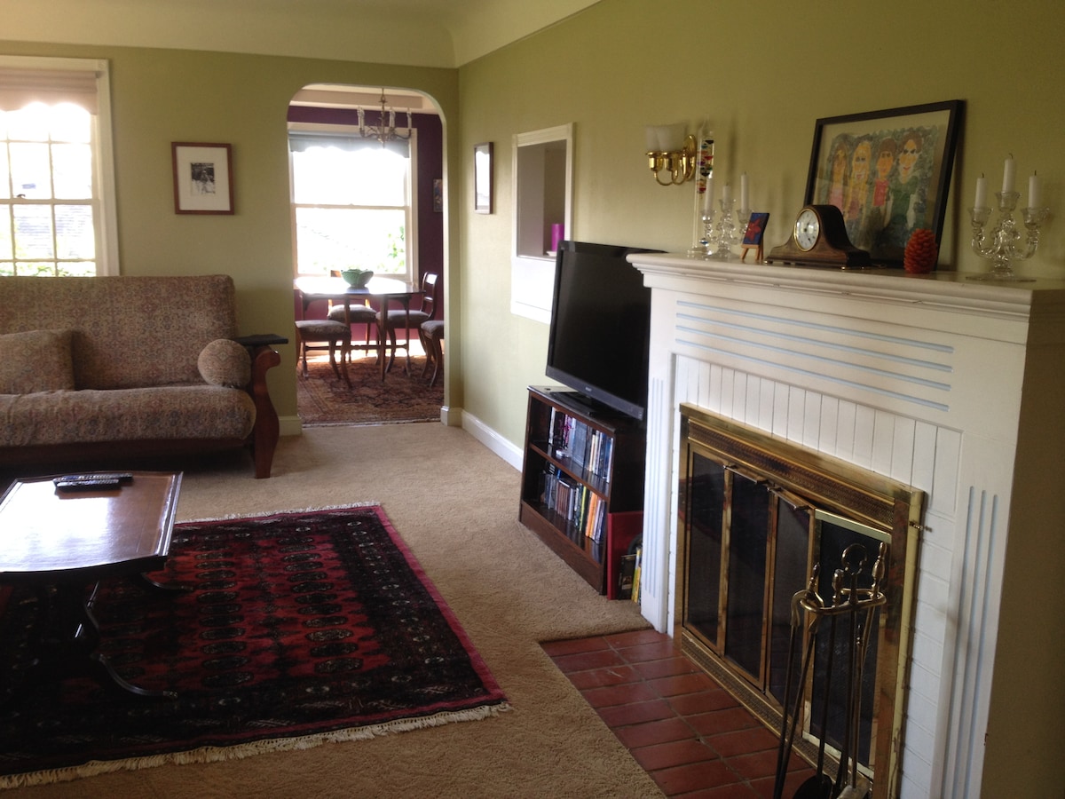 The living area is furnished with a patterned sofa and a wooden coffee table. A brick fireplace is positioned next to a bookcase. A dining area with a table and chairs can be seen through an archway, accompanied by a colorful wall art piece above the fireplace.