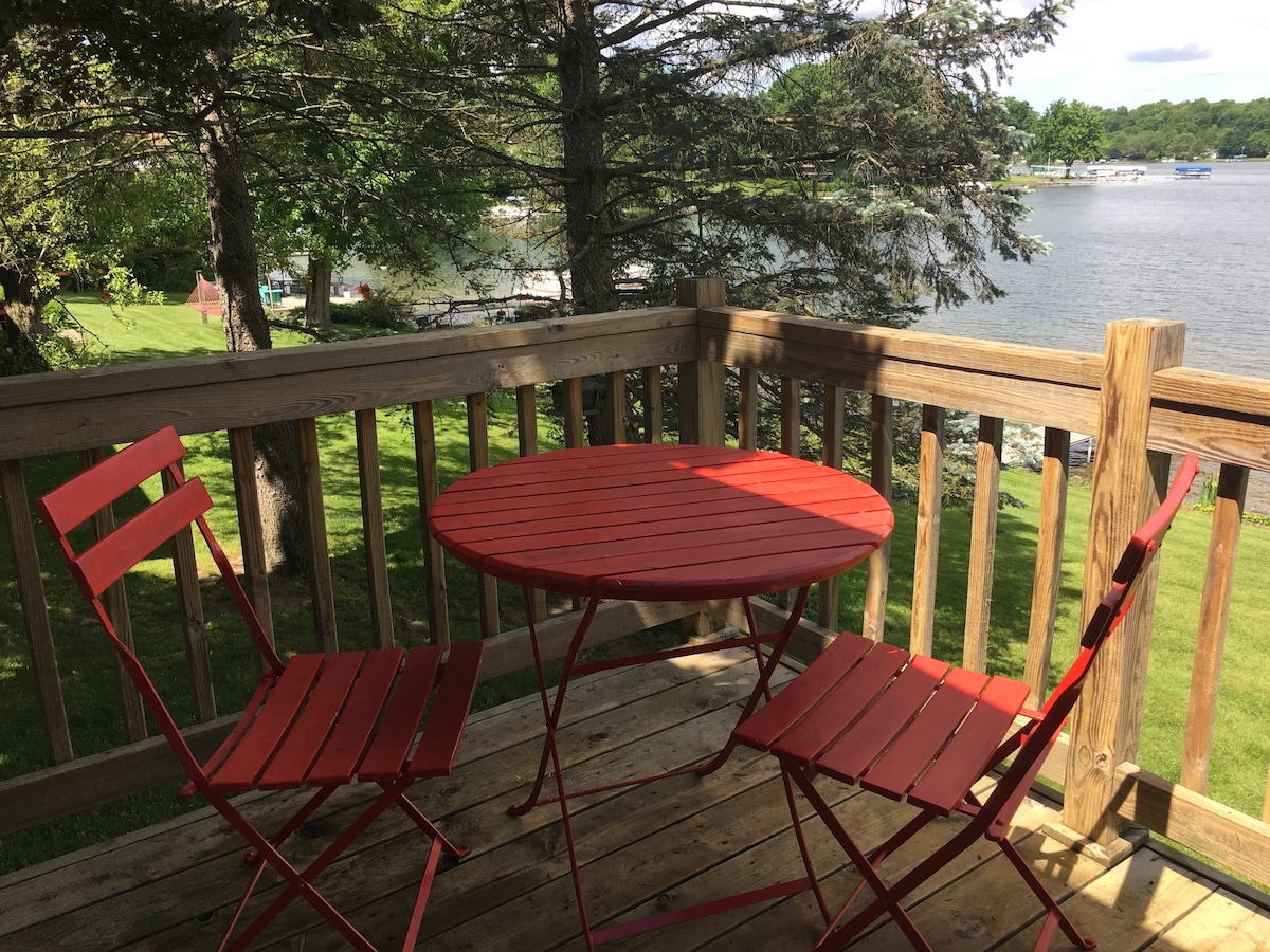 A wooden deck features a round red table surrounded by two matching folding chairs. Lush green grass extends to the water's edge, with several boats visible on the lake in the background, framed by tall trees providing shade.
