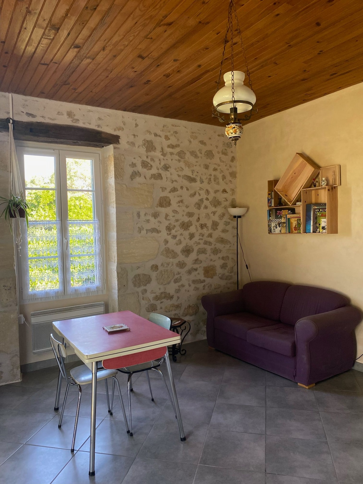 A cozy corner is showcased with a purple sofa and a small dining table surrounded by metal chairs. A window allows natural light to illuminate the space, while a unique bookshelf displays various items against the textured stone wall.