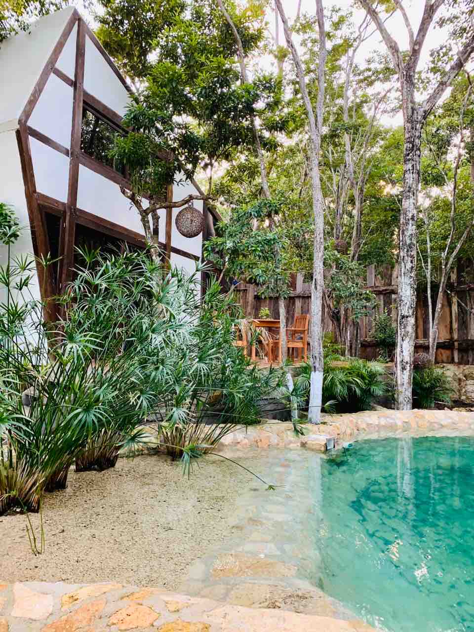 A serene natural pool is surrounded by lush greenery, with tall trees and tropical plants enhancing the tranquil environment. In the background, a uniquely designed house features wooden accents. A small outdoor seating area with a wooden table is visible, offering a peaceful spot to relax.