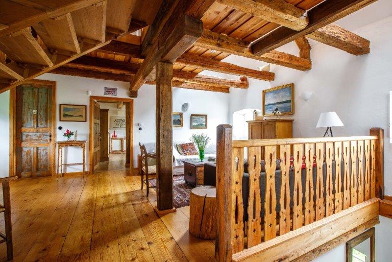The interior space features exposed wooden beams and a warm wooden floor. A seating area is arranged around a coffee table, accompanied by framed artwork on the walls. Natural light floods the room through large windows, while a rustic wooden railing borders the upper level.
