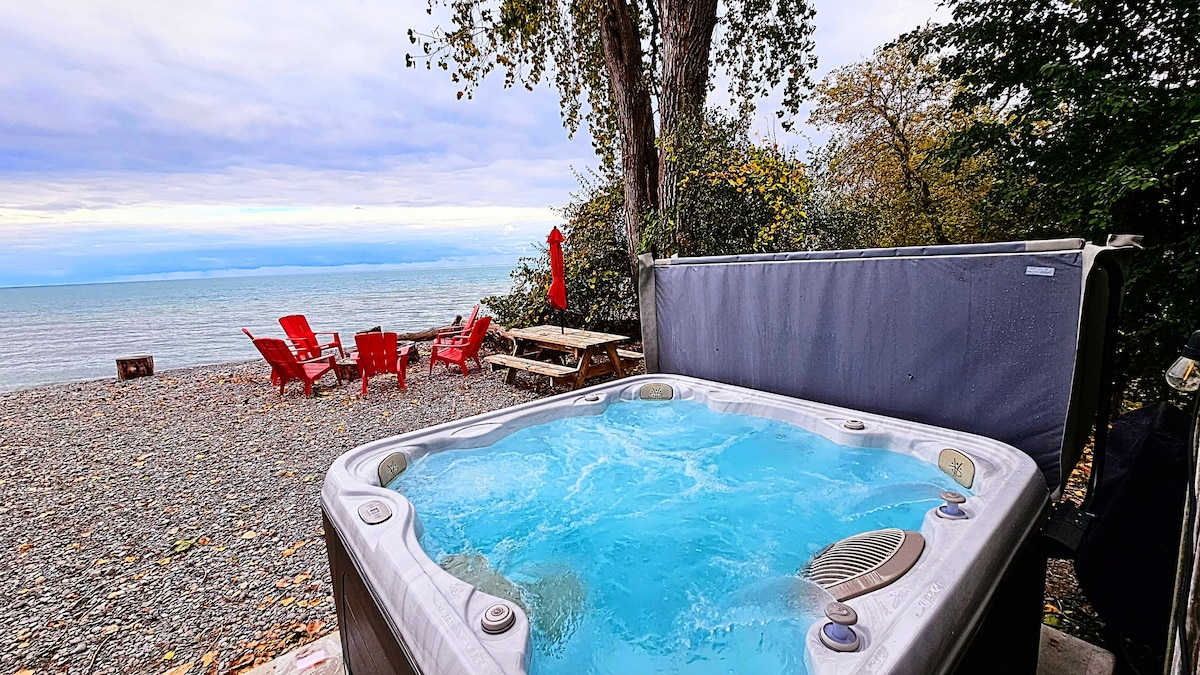 A hot tub is positioned near a private shoreline, offering a view of the lake. Adirondack chairs in vibrant red are arranged on a pebbled area, alongside a picnic table shaded by trees, enhancing the outdoor relaxation space.