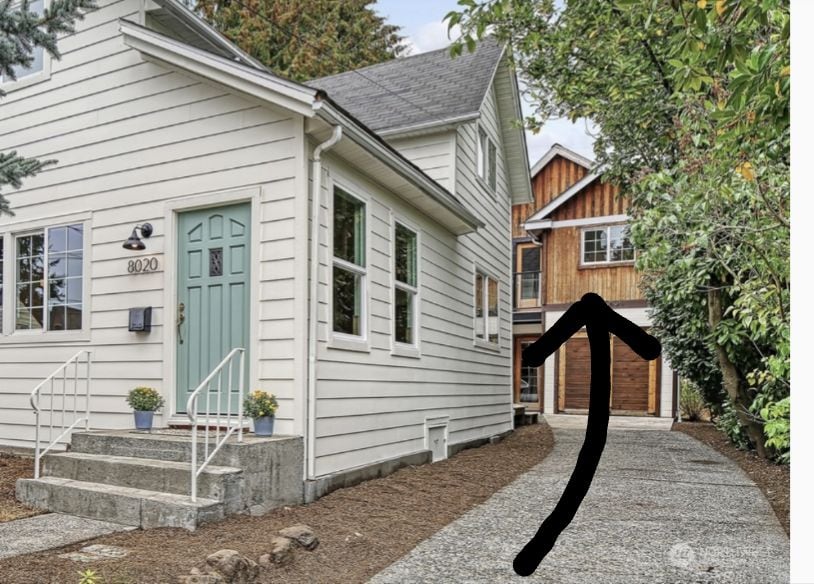A charming white exterior of a farmhouse is visible, with a turquoise door and neatly arranged flower pots. A pebbled driveway leads towards a wooden structure, suggesting an inviting path to the separate unit in the backyard surrounded by greenery.
