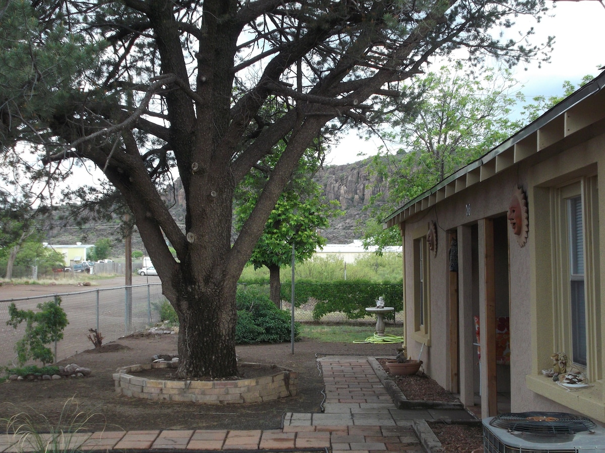 A large tree provides shade in the yard, accompanied by lush greenery and distant mountainous terrain. The adobe home is partially visible, featuring a patio area with stone paving. Soft natural light filters through the leaves, enhancing the tranquility of the outdoor space.