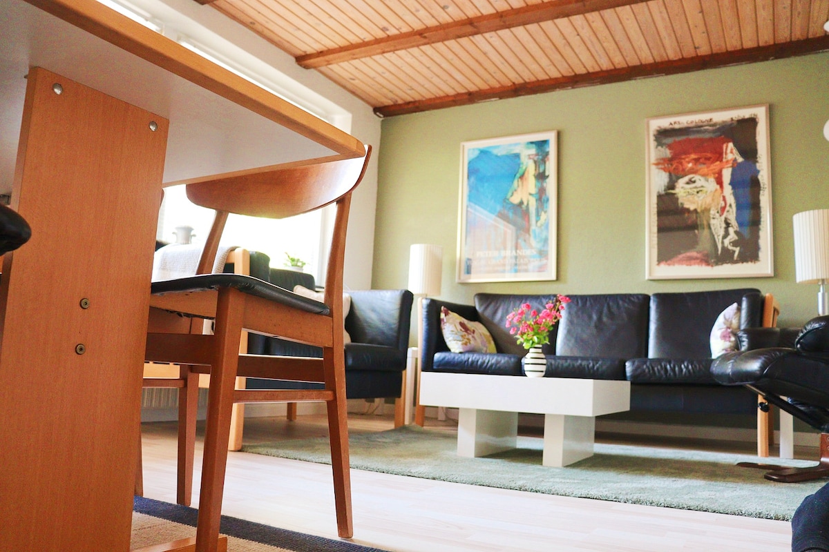 A spacious living area features two black leather sofas facing a central coffee table. A wooden dining table and chair are positioned nearby, complemented by colorful wall art and a vase of flowers adorning the table.