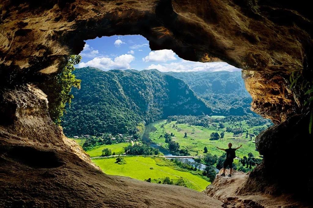 A natural cave opening frames a breathtaking view of lush green hills and valleys. The landscape features rolling hills, a winding river, and scattered trees beneath a clear blue sky. A figure stands within the cave, adding a sense of scale to the towering rock formations.