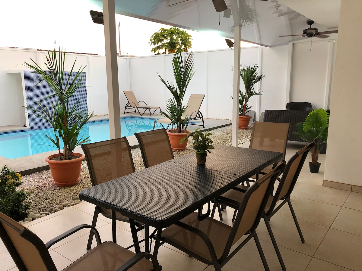 An outdoor dining area is set up with a large table and several chairs, surrounded by potted plants. A swimming pool glimmers in the background, while lounge chairs are positioned nearby, providing a relaxed atmosphere.