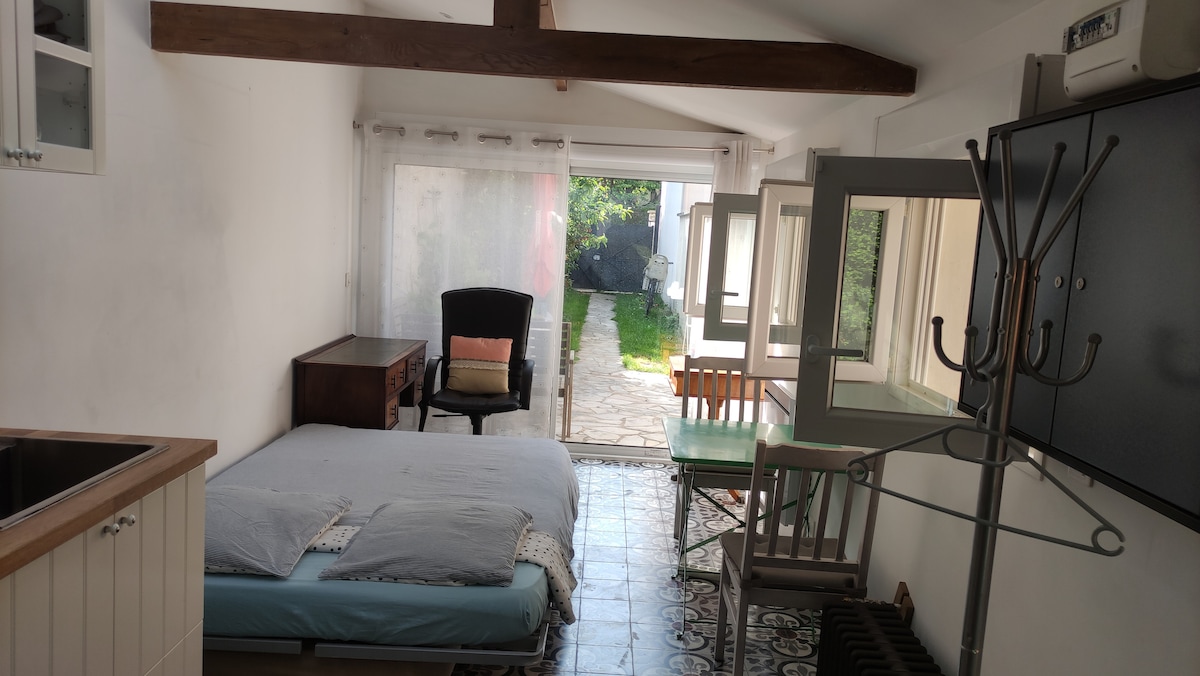 A bright room is illuminated by natural light from large windows, featuring a spacious bed with soft linens. A small dining table is positioned nearby, with a chair and a coat stand visible. A vintage armchair adds character to the space, which is complemented by patterned flooring.