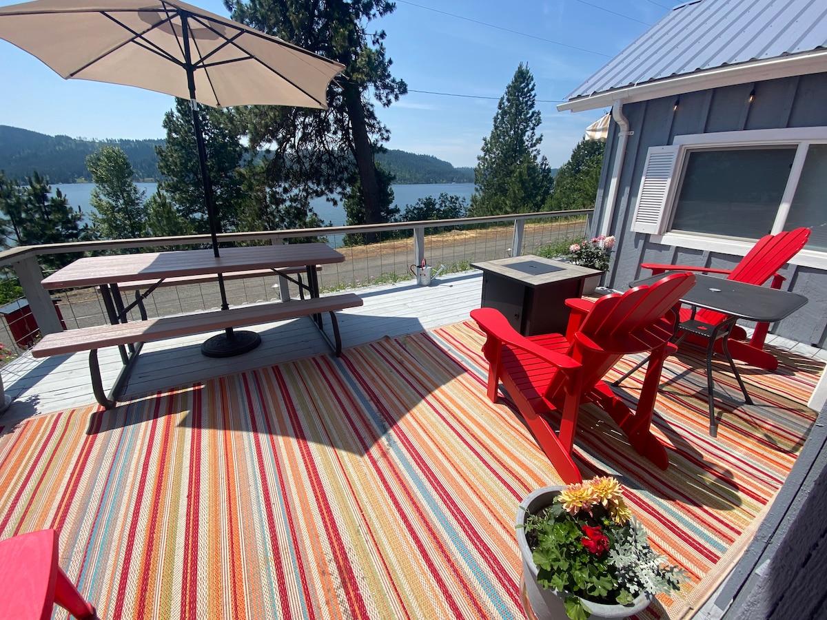 An outdoor deck area features a large striped rug and is furnished with red Adirondack chairs and a picnic table. An umbrella provides shade, overlooking a scenic view of the lake and surrounding trees, enhancing the relaxing atmosphere of the space.