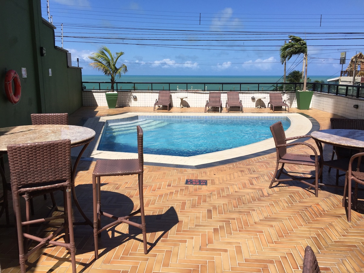 A sunlit pool area features a clear blue swimming pool surrounded by laid-back lounge chairs. Tables with woven seats provide space for relaxation, all set against a backdrop of ocean views and blue skies.