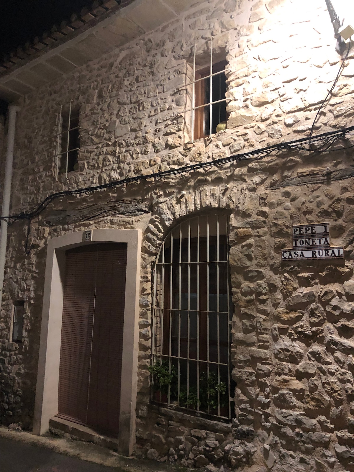 The stone façade of a rustic house is illuminated softly at night. A wooden door with slatted blinds is framed by an arched window with decorative bars. The house displays a sign reading 'Pepe y Tonita Casa Rural', highlighting its welcoming charm.