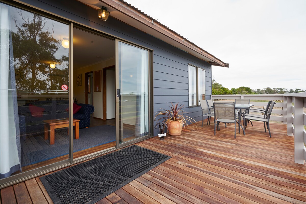 A large wooden deck is shown, featuring a seating area with a table and chairs. Sliding glass doors lead into a spacious lounge area, while greenery is visible in the background. Natural light highlights the deck's surface and the surrounding landscape.