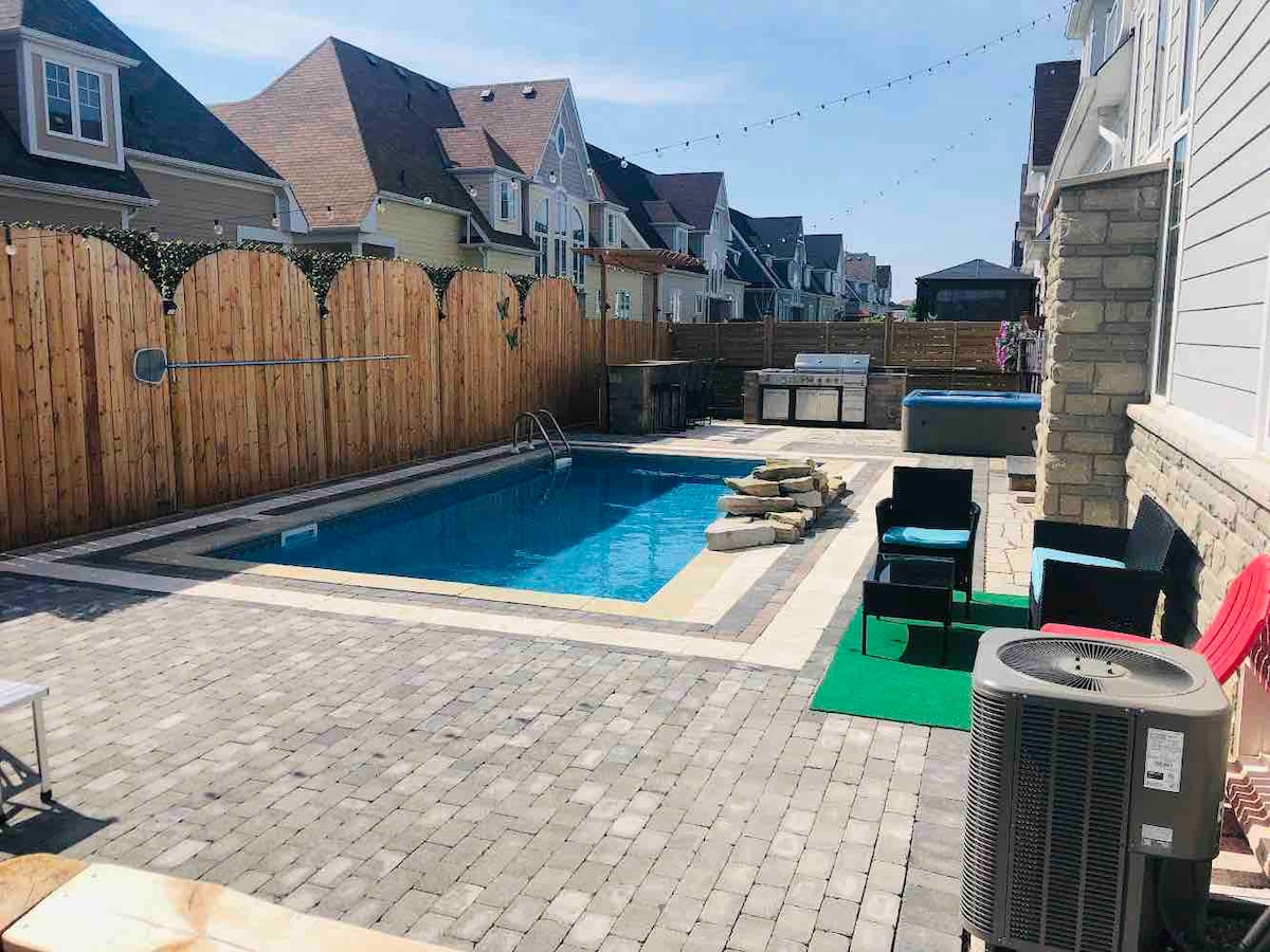 An outdoor space features a rectangular in-ground heated pool surrounded by paved decking. A hot tub is visible in the background, alongside a BBQ area and a wooden fence providing privacy. Seating is arranged with black chairs and loungers set around the pool.