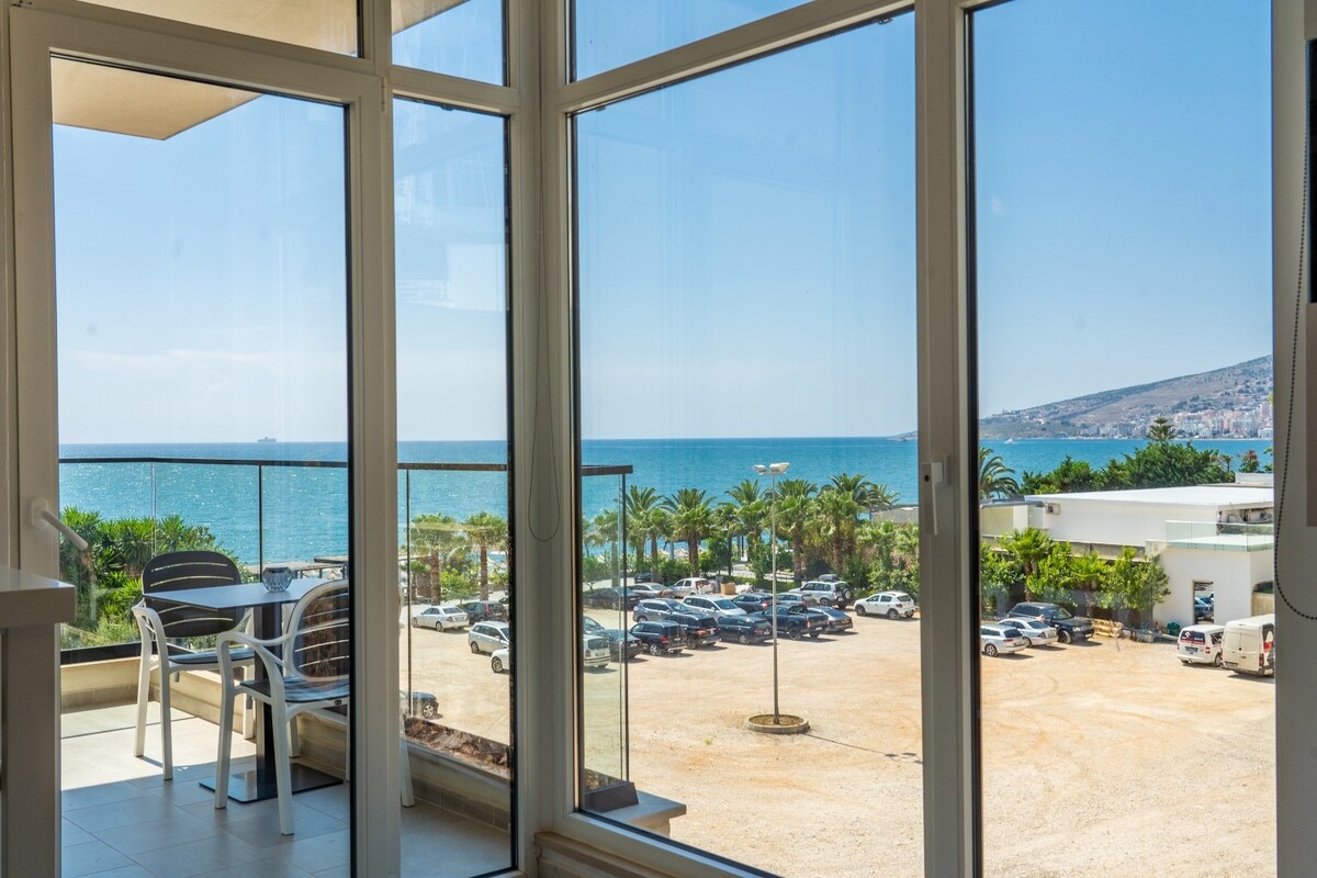 A panoramic view of the tranquil sea and distant hills is framed by large glass doors. A small balcony with two chairs is visible, inviting relaxation. Below, a parking area is filled with vehicles, bordered by palms that enhance the coastal setting.