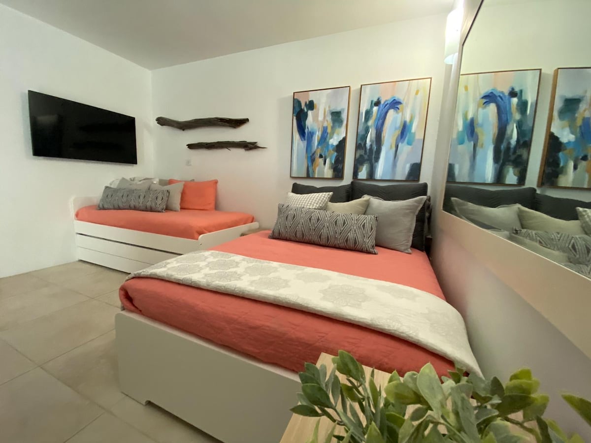 A compact sleeping area features a full bed and a pull-out single bed, both adorned with colorful bedding and decorative pillows. A large mirror reflects the contemporary artwork on the wall, while a flat-screen TV is mounted on the opposite wall.