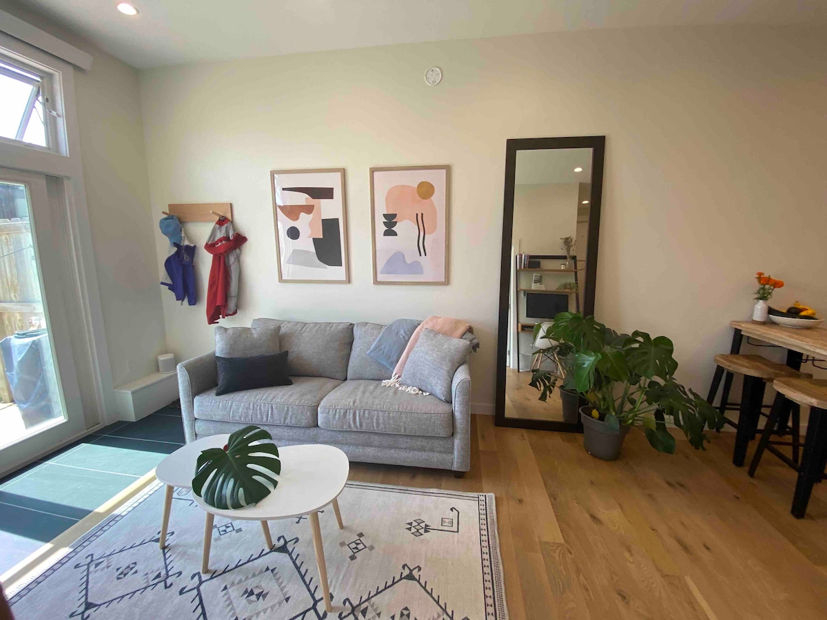 A living room features a cozy gray couch positioned beside a small round coffee table on a patterned area rug. Colorful wall art hangs above the couch, and a large mirror is located nearby. A green potted plant adds a touch of nature to the space.