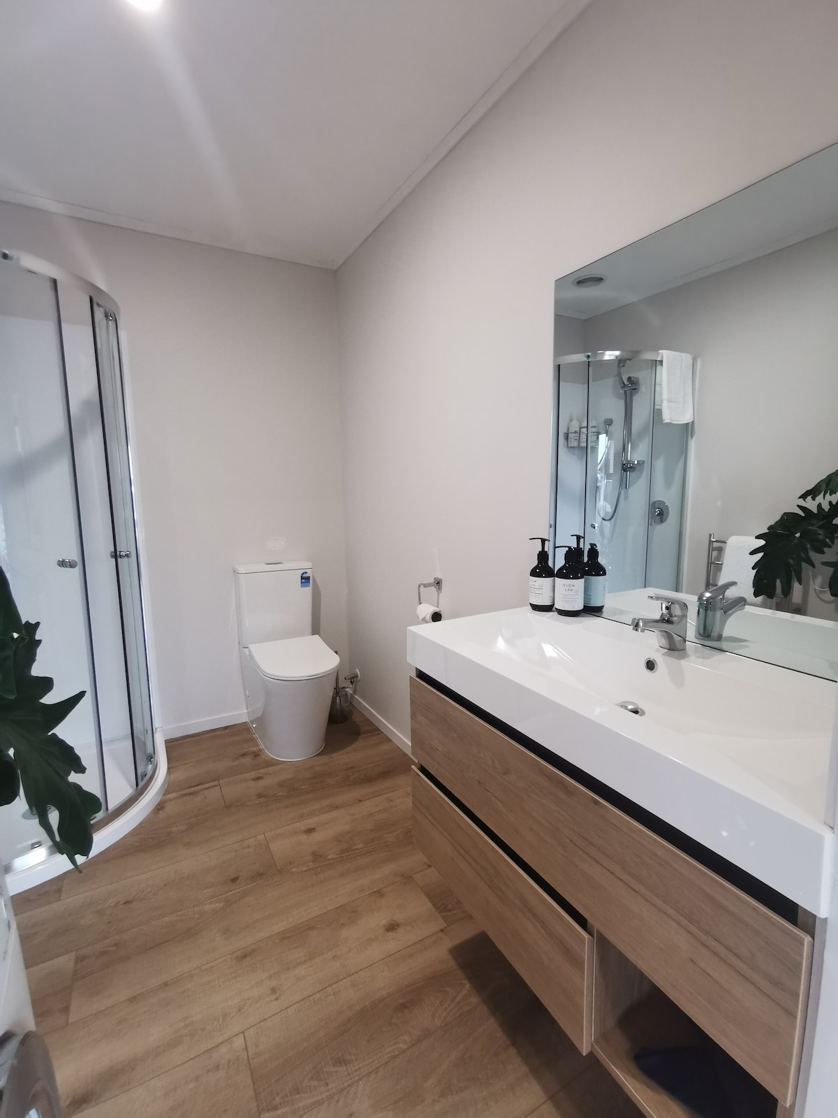 The bathroom features a modern design with a glass shower enclosure and wood-textured cabinetry beneath a sleek white countertop. A mirror above the sink reflects the space, while neatly arranged toiletries are displayed on the counter. Natural light enhances the clean, minimalist atmosphere.