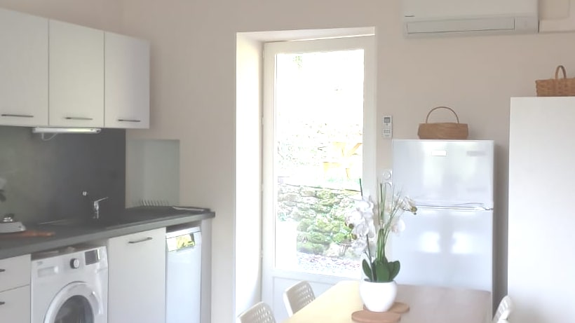 A clean and modern kitchen area features sleek white cabinetry and a dark countertop. A dining table with light-colored chairs is positioned near a window, which offers views of the surrounding greenery. Appliances, including a refrigerator and washing machine, are clearly visible.