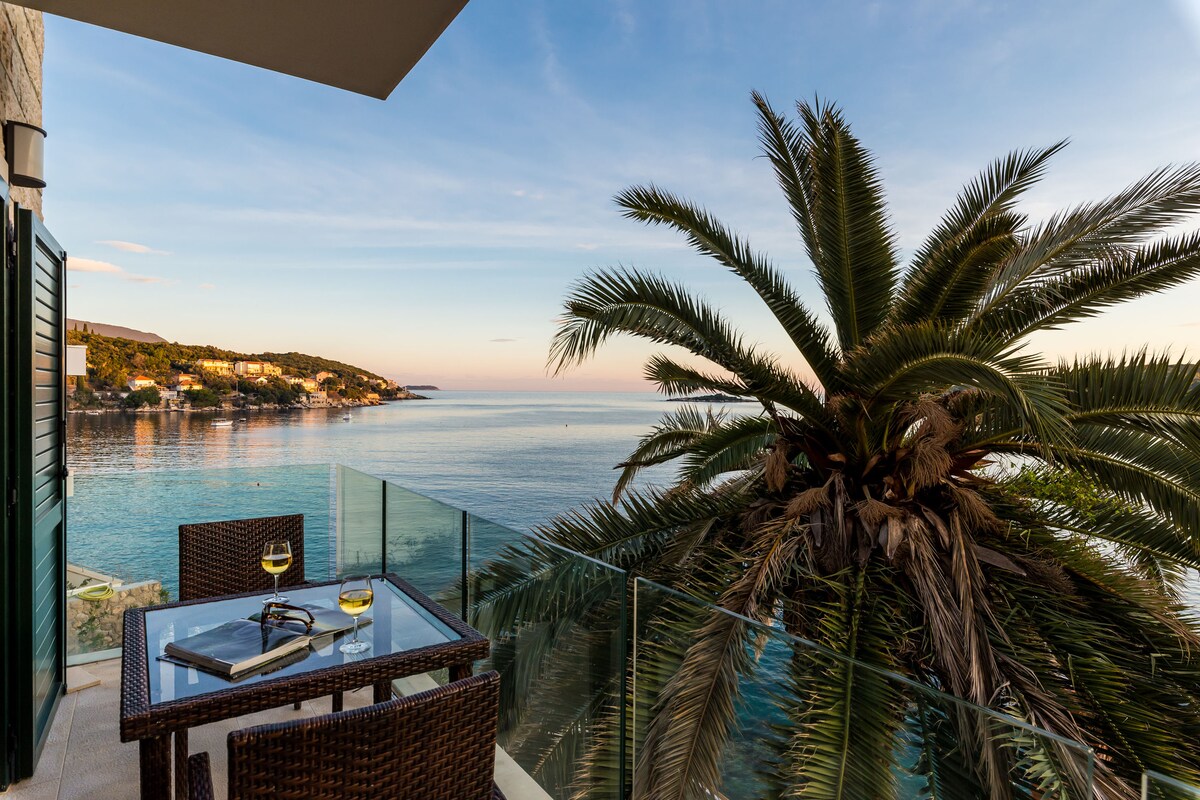 A balcony offers a serene space with a glass railing, featuring a wicker table and two chairs set against a backdrop of palm trees. The sea reflects golden hues under a clear sky, creating a calming coastal view.
