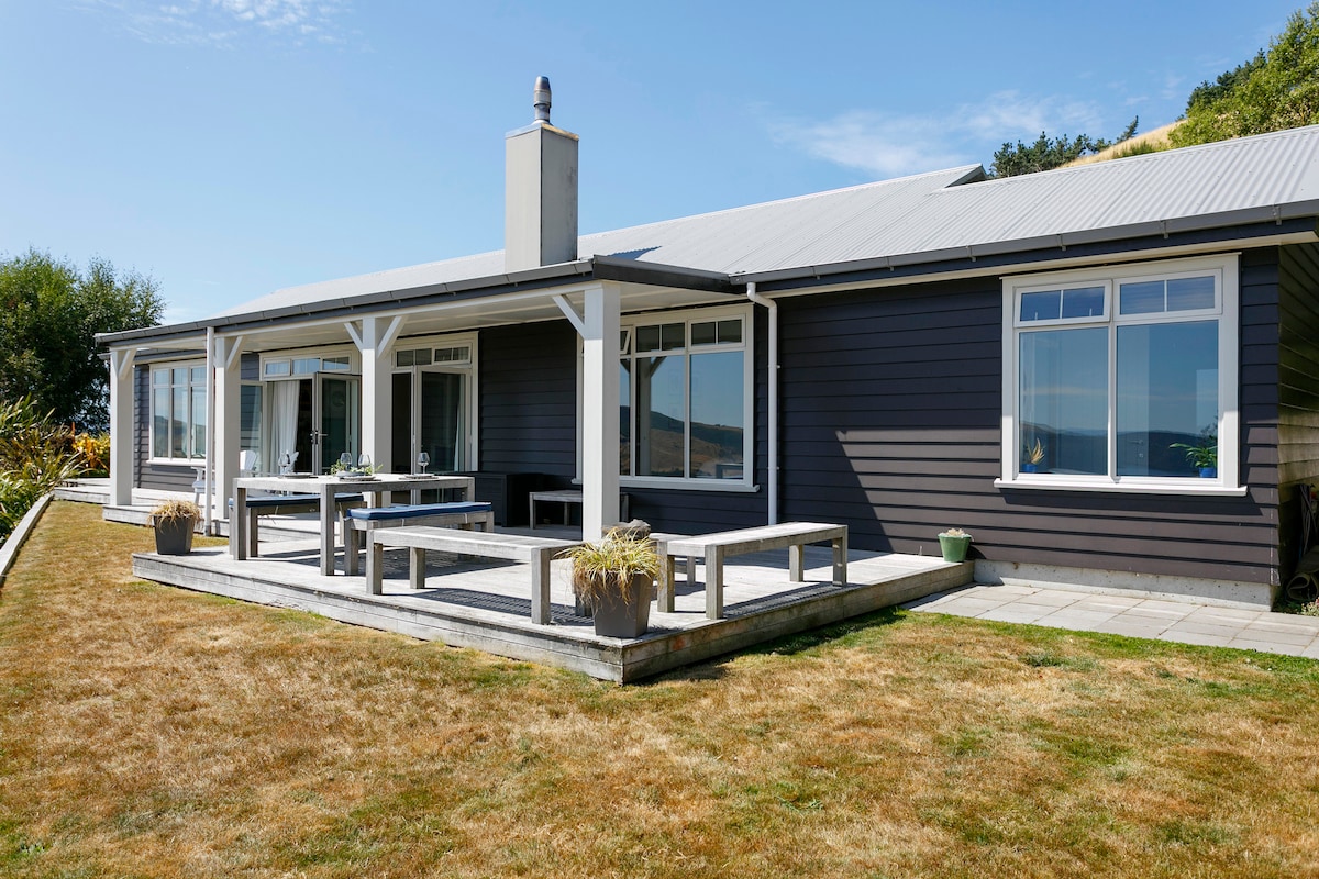 The exterior of the house features a large deck area with wooden seating arrangements. Glass sliding doors provide a view of the interior, while the surrounding lawn adds to the spaciousness. A clear blue sky complements the design, enhancing the inviting ambiance.