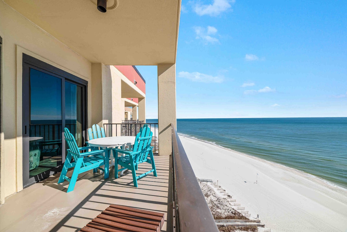 The spacious balcony features colorful adirondack chairs and a table, overlooking the expansive beach and emerald waters. The coastline stretches along the horizon, while the clear blue sky enhances the serene atmosphere of the outdoor space.