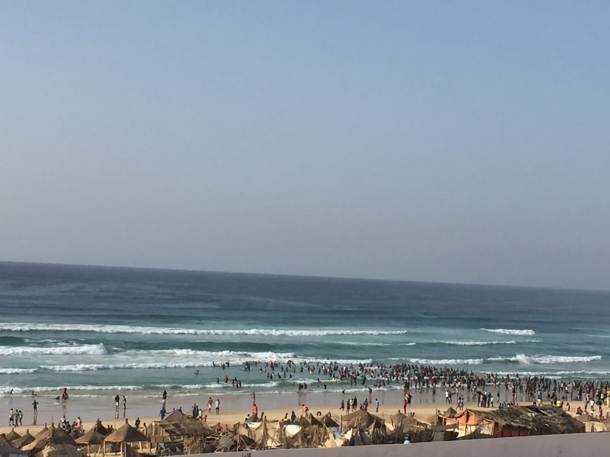 A wide view of a beach scene shows numerous people enjoying the water along a sandy shoreline. Gentle waves lap at the beach, while sun loungers and umbrellas are visible in the foreground. The vast ocean stretches toward a clear sky.