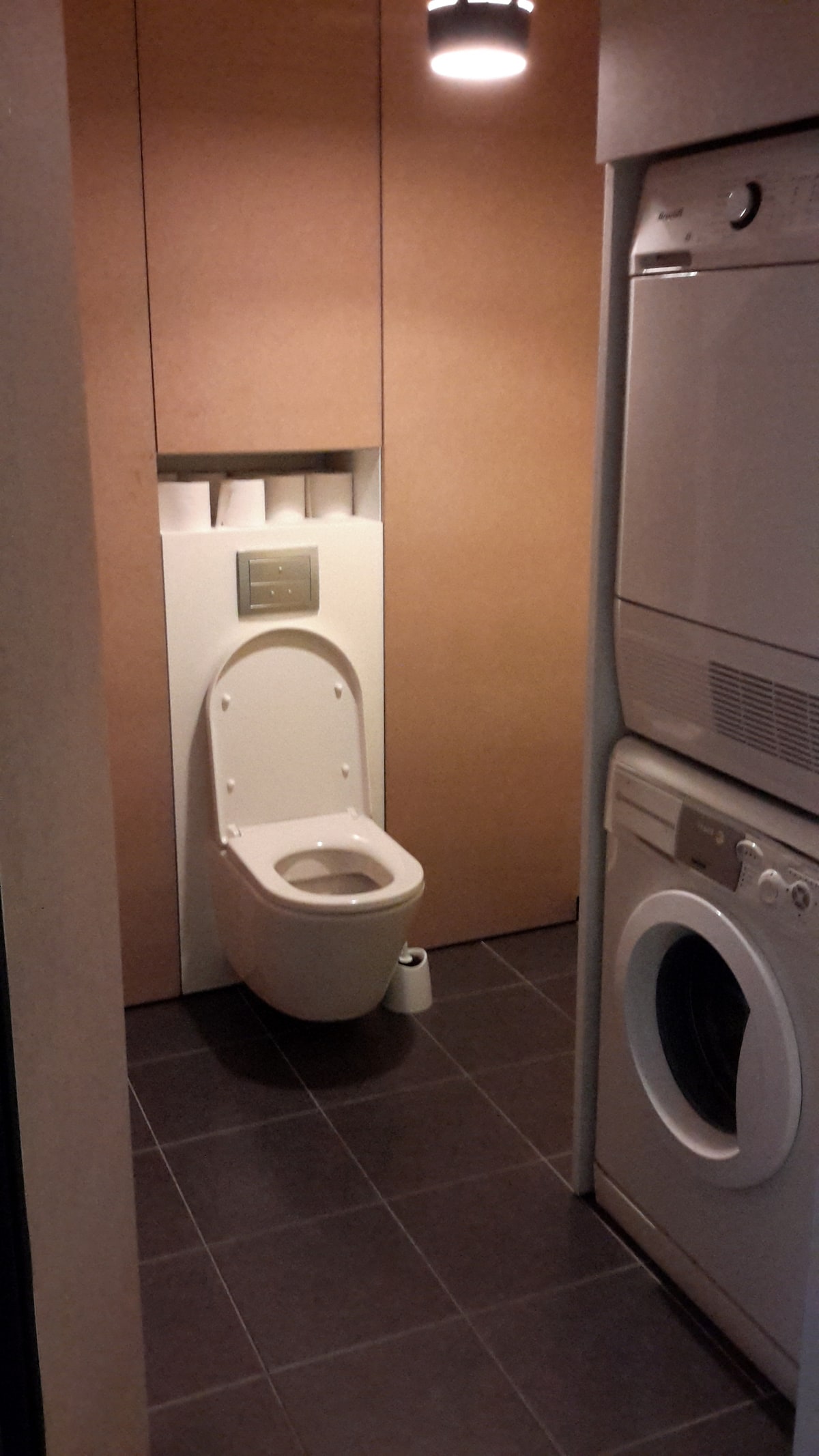 A compact bathroom space features a modern, wall-mounted toilet positioned against a backdrop of textured panels. Adjacent to the toilet, a stacked washer and dryer are located, providing convenience within the area. The floor is tiled in dark tones, complementing the neutral walls.