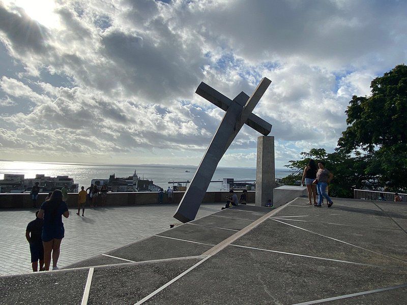 A large, modern sculpture of a cross is prominently placed on a terrace overlooking the ocean. The scene features visitors enjoying the view against a backdrop of cloudy skies and the horizon. The area is paved with geometric patterns, enhancing the outdoor space.