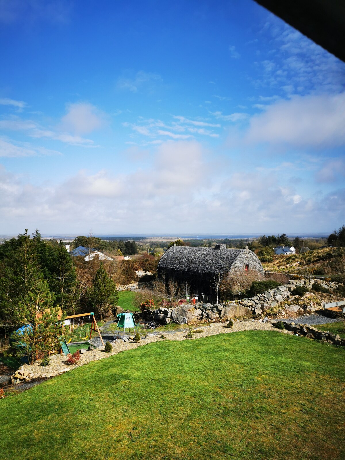 A spacious garden area is featured, showcasing lush green grass and rocky borders. In the background, a traditional stone barn is visible under a blue sky. Nearby, play equipment can be seen, suggesting a family-friendly environment surrounded by scenic rural landscape.