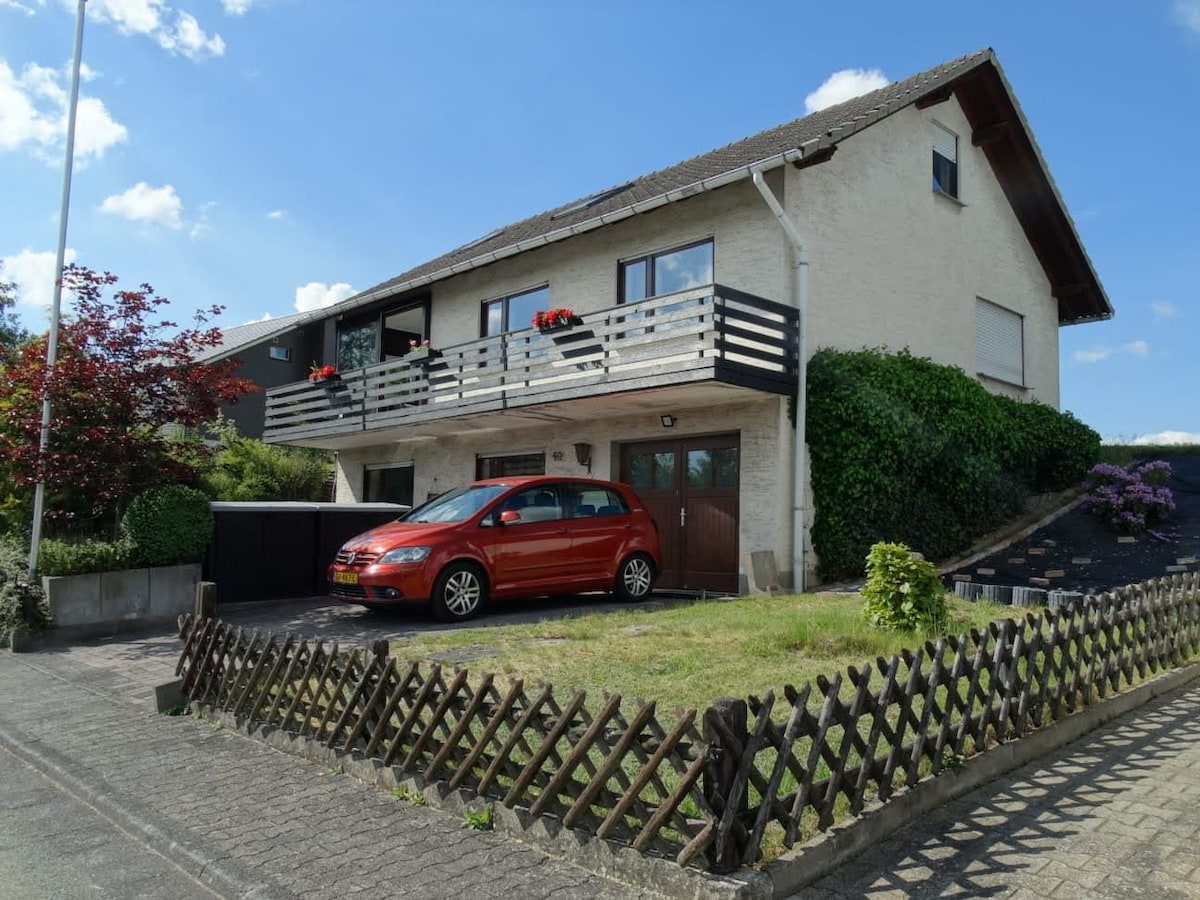 A two-story house is depicted, featuring a front garden with neatly trimmed grass and a wooden fence. A red car is parked in the driveway. Flower boxes adorn the balcony, and a pathway leads to the entrance, surrounded by greenery and blooming plants.