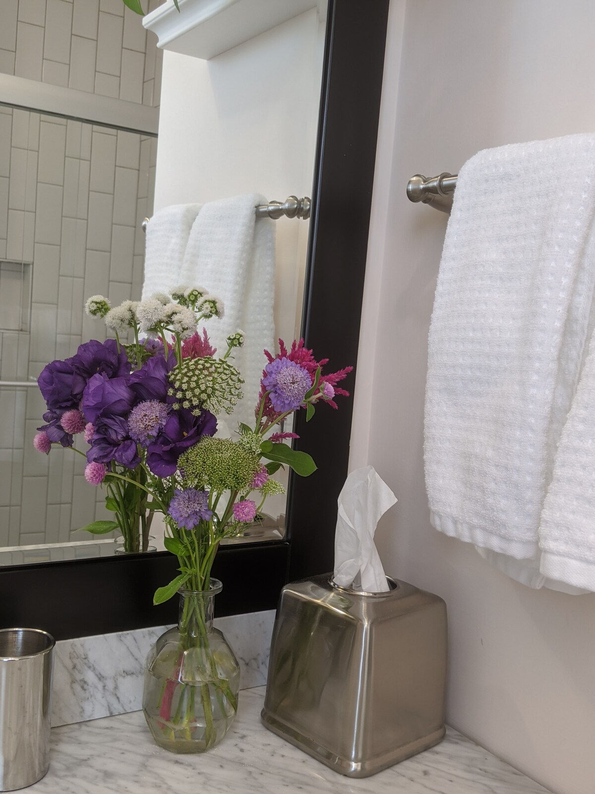 A modern bathroom features a marble countertop with a stainless steel tissue box and a polished metal cup. White towels hang neatly on a bar beside a mirror, reflecting a vase of vibrant purple and white flowers, adding a touch of color to the space.
