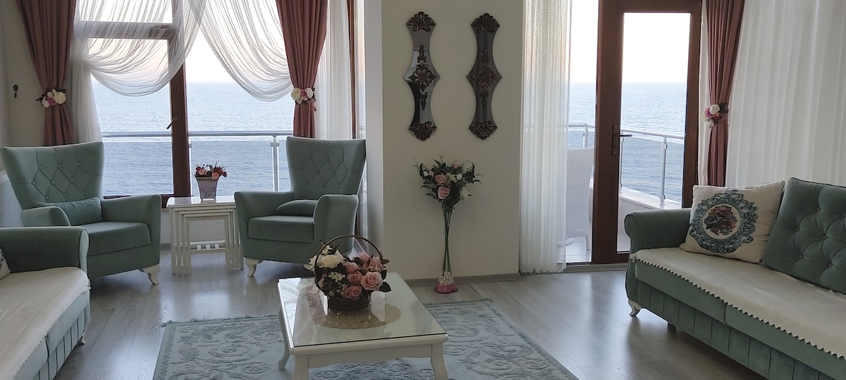 A light-filled living area features two soft blue armchairs and a comfortable white sofa. Decorative mirrors adorn the wall, while a vase of flowers rests on a glass coffee table. Large windows showcase unobstructed views of the sea, enhancing the serene environment.
