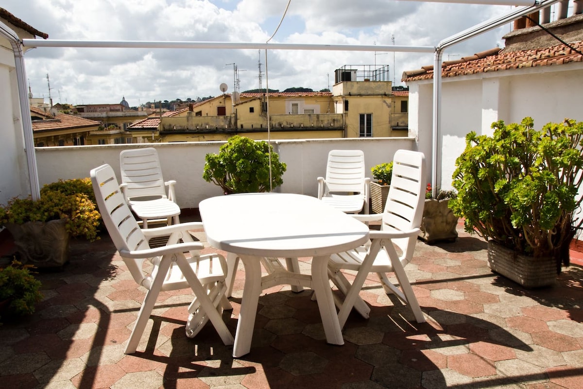 A private terrace is presented with a white outdoor table and six matching chairs, surrounded by potted plants. The view captures a portion of the Roman skyline, providing a bright and open feel under a partly cloudy sky.