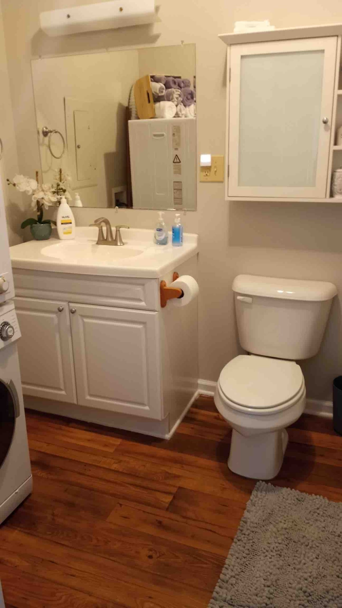 A compact bathroom is presented, featuring a white countertop with a sink and ample storage cabinets. A toilet is situated adjacent to the sink. Warm wooden flooring complements the space, while a soft area rug adds comfort. A mirror reflects natural light, enhancing the bright atmosphere.