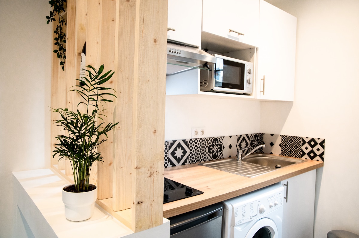 The kitchen showcases a modern design with wood accents and black and white patterned tiles. A compact washing machine is situated beside a stainless steel sink. A microwave and cooking stove are well-integrated into the cabinetry, while a potted plant adds a touch of greenery.