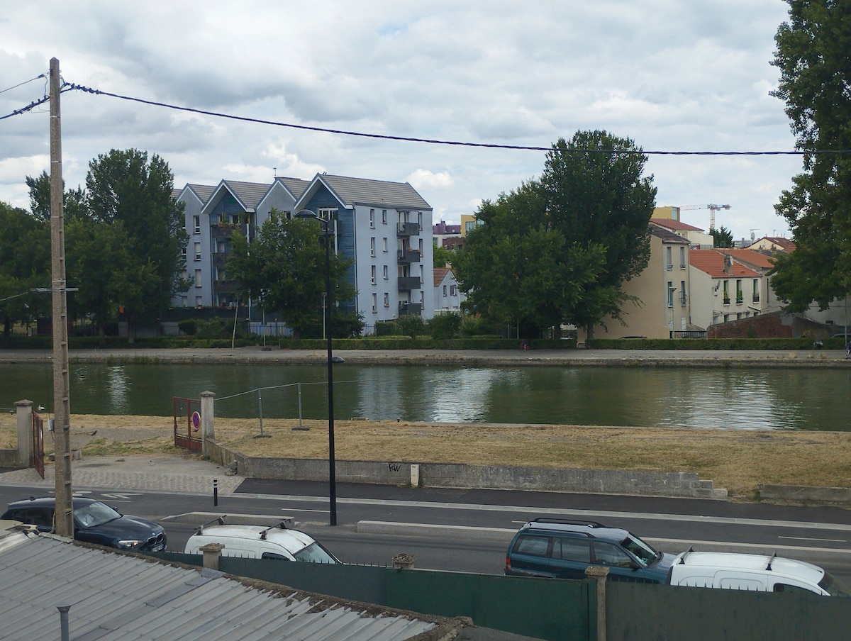 A view features a calm canal bordered by trees, with various residential buildings visible across the water. The landscape includes a grassy area and a road lined with parked cars, contributing to the tranquil setting.