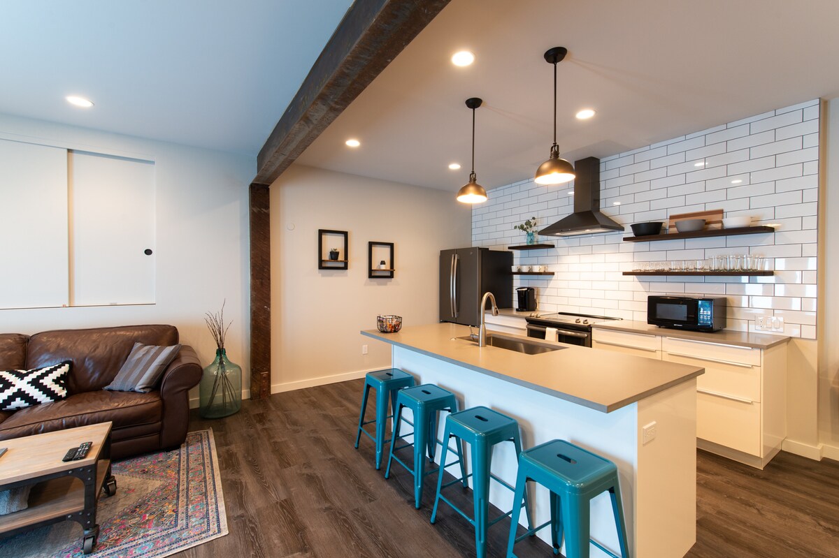 The kitchen area features a modern design with a large island equipped with blue stools. A variety of kitchen appliances, including a black refrigerator and sleek range hood, are visible. A comfortable brown sofa sits in the living space, complemented by decorative prints on the walls.