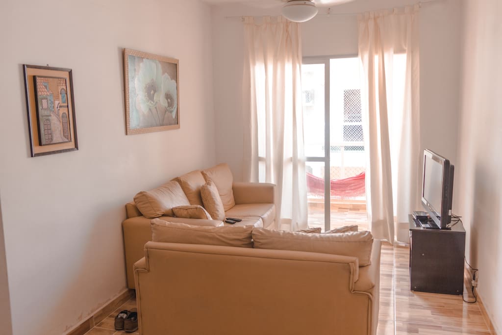 Sala ampla com sofás, tv, mesa de jantar, cortina e sacada com rede para descanso