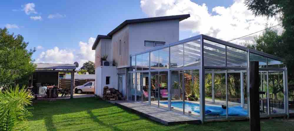 A modern two-story house is surrounded by lush greenery, featuring large glass walls that create an open feel. The patio includes a sheltered barbecue area and a heated pool, offering a seamless indoor-outdoor connection. Bright skies and natural light enhance the overall ambiance.