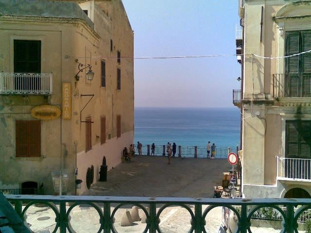 A view from a balcony frames the serene sea, with gentle waves meeting the shore in the distance. Nearby, a square is visible, where several people gather. The architecture displays traditional Italian buildings, featuring balconies and large windows, creating a charming coastal ambiance.