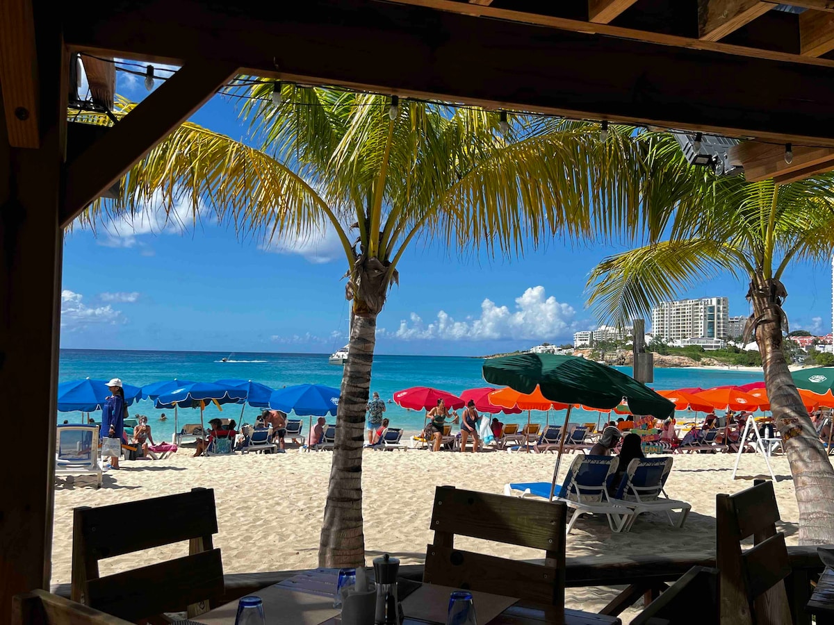 A view from beneath a wooden structure reveals a sandy beach lined with colorful umbrellas. Palm trees create shade over loungers, while gentle waves lap at the shore in the background. The blue sky is dotted with a few clouds, enhancing the coastal atmosphere.