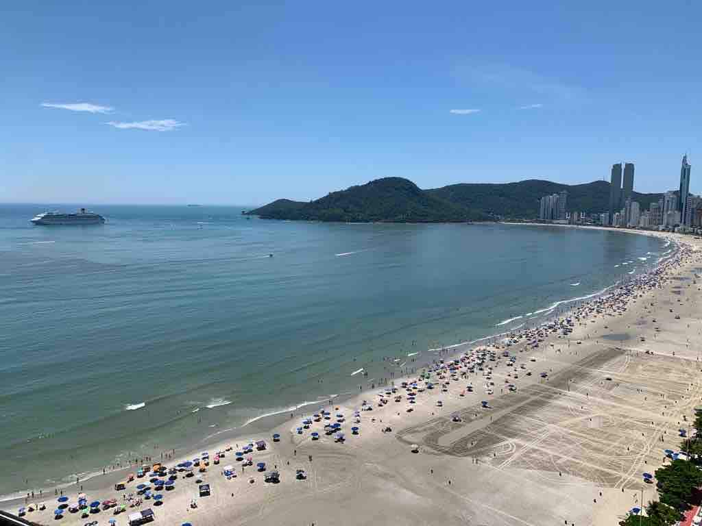 The beach view stretches across the image, showcasing golden sand lined with colorful umbrellas. Gentle waves lap at the shore, while a distant coastal mountain adds to the scenic backdrop. The vibrant atmosphere is complemented by a few boats visible in the water.
